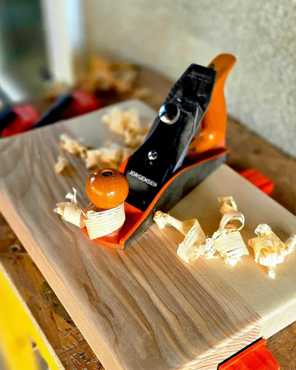 Rabot à main et copeaux posés sur une planche de bois clair, sur l'établi