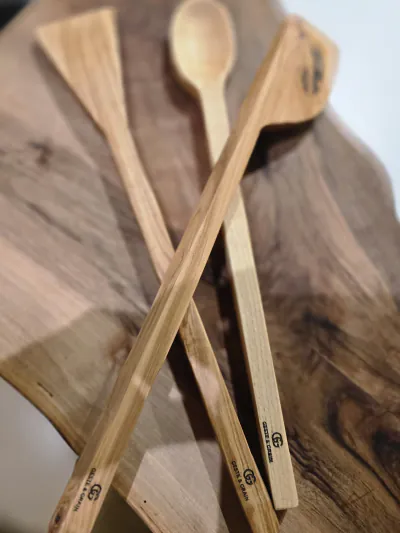 Deux spatules et une cuillère en bois clair gravées Geste & Grain, posées sur une planche de bois sombre